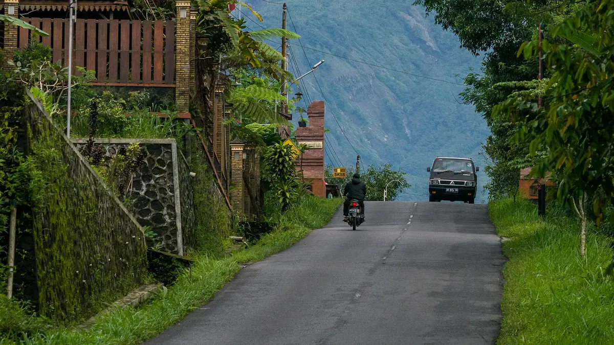 Jalan desa menanjak di Kalurahan Kepuharjo, Cangkringan, dengan pengendara motor dan mobil bak di depan lereng Merapi