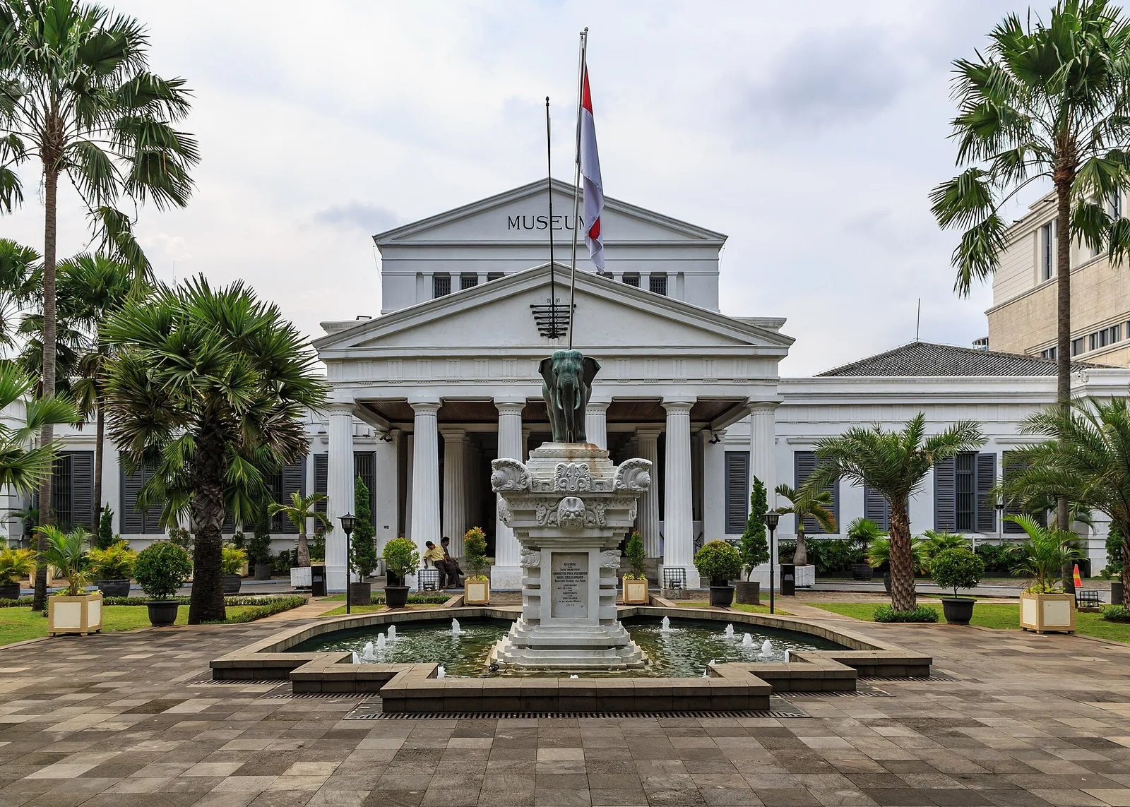 Patung gajah perunggu dan kolam air mancur di halaman depan Museum Nasional Indonesia, dengan gedung berpilar bergaya kolonial di belakangnya