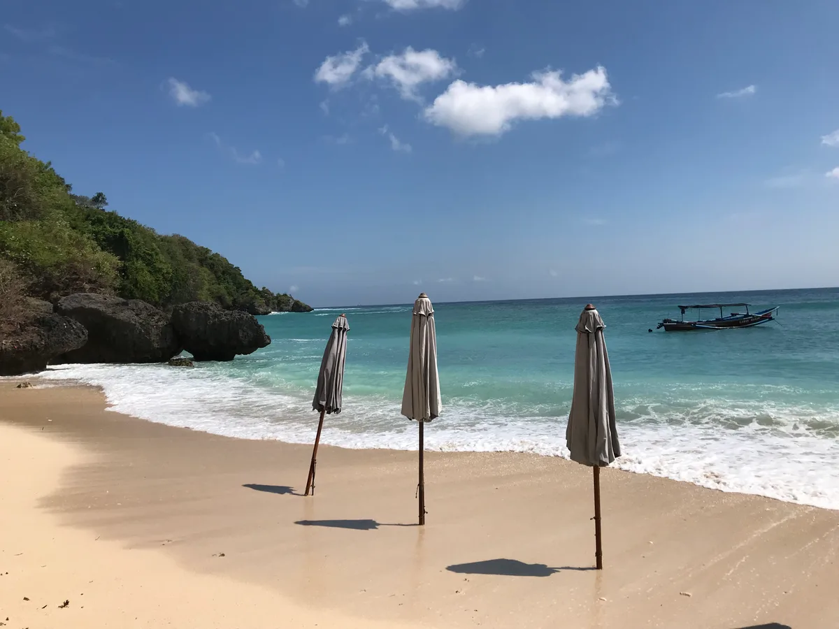 Tiga payung pantai tertutup berdiri di pasir Pantai Padang Padang menghadap air toska dan perahu kecil