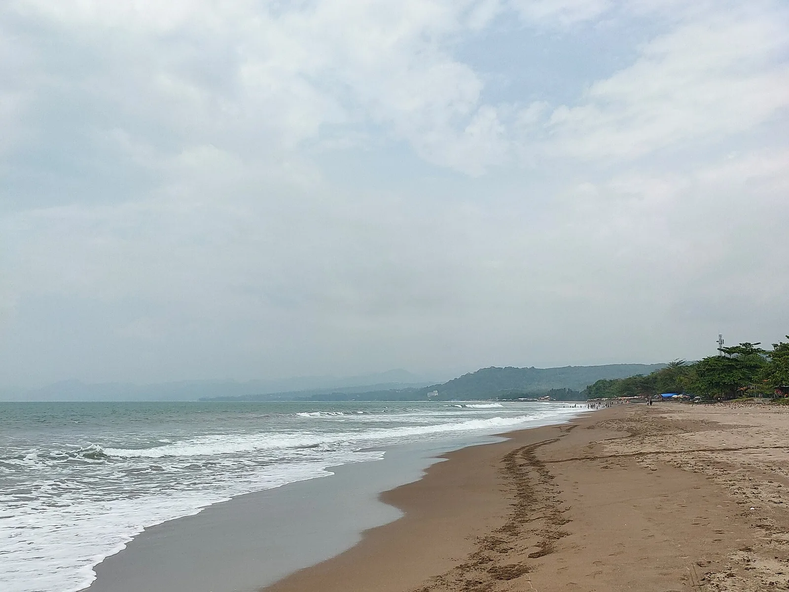 Garis Pantai Citepus yang panjang dengan ombak putih, pasir kecokelatan, dan deretan warung bertenda di kejauhan menuju perbukitan