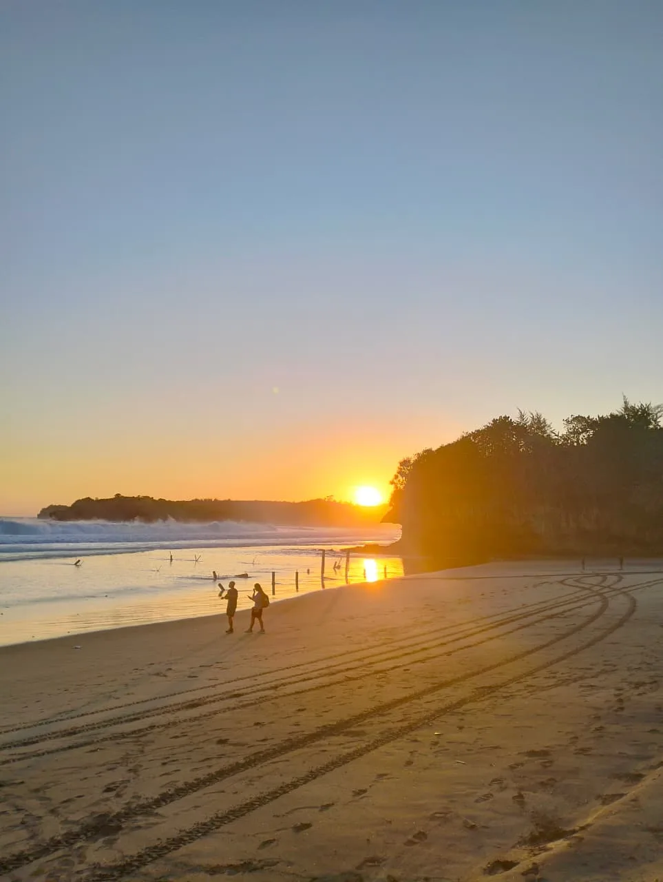 Matahari terbenam keemasan di Pantai Serang Blitar dengan tanjung berhutan di sisi kanan dan jejak roda di pasir basah