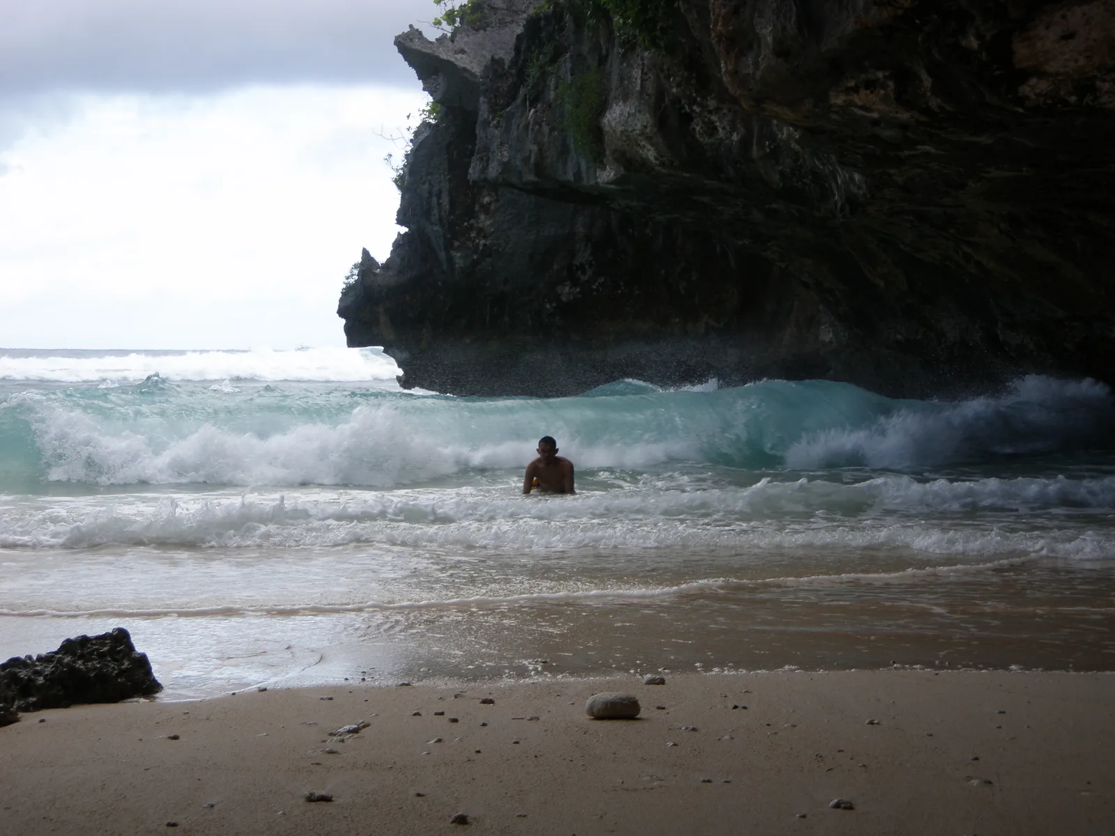 Ombak pecah di depan tebing karang menjorok di Pantai Suluban dengan seorang perenang di antara buih