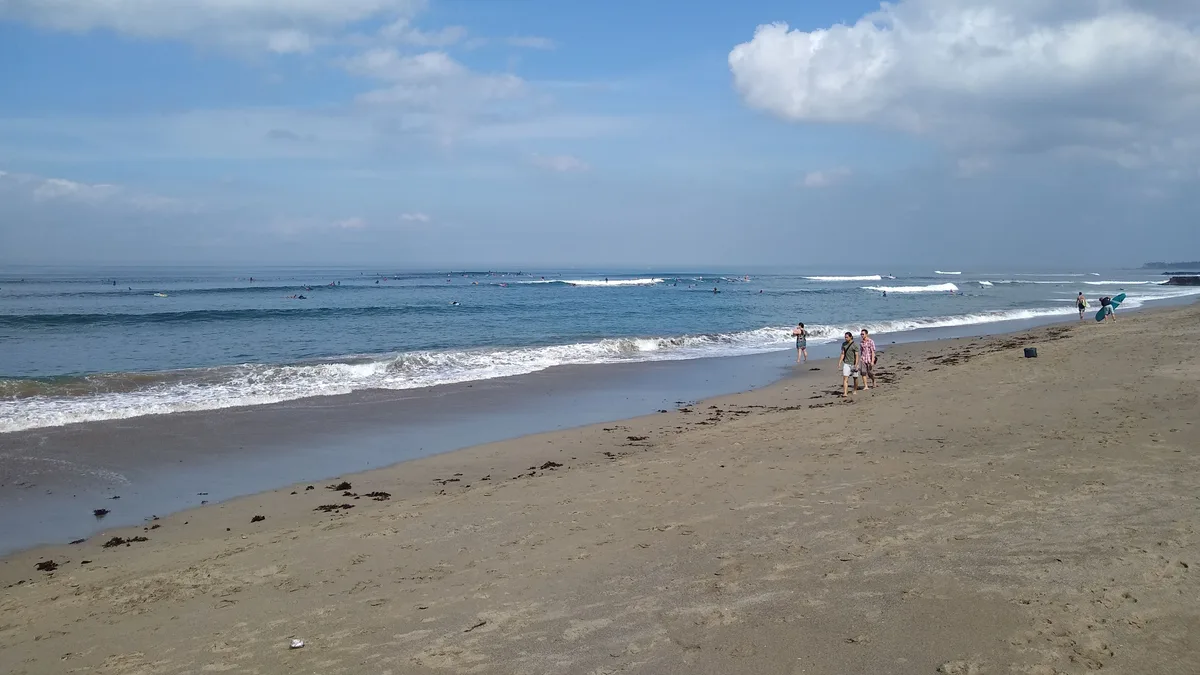 Ombak bergulung di Pantai Canggu dengan pasir gelap dan beberapa peselancar di kejauhan