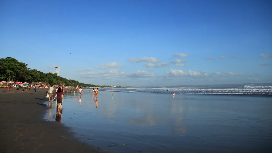 Garis Pantai Kuta yang panjang dengan pengunjung berjalan di pasir basah dan ombak kecil di kejauhan
