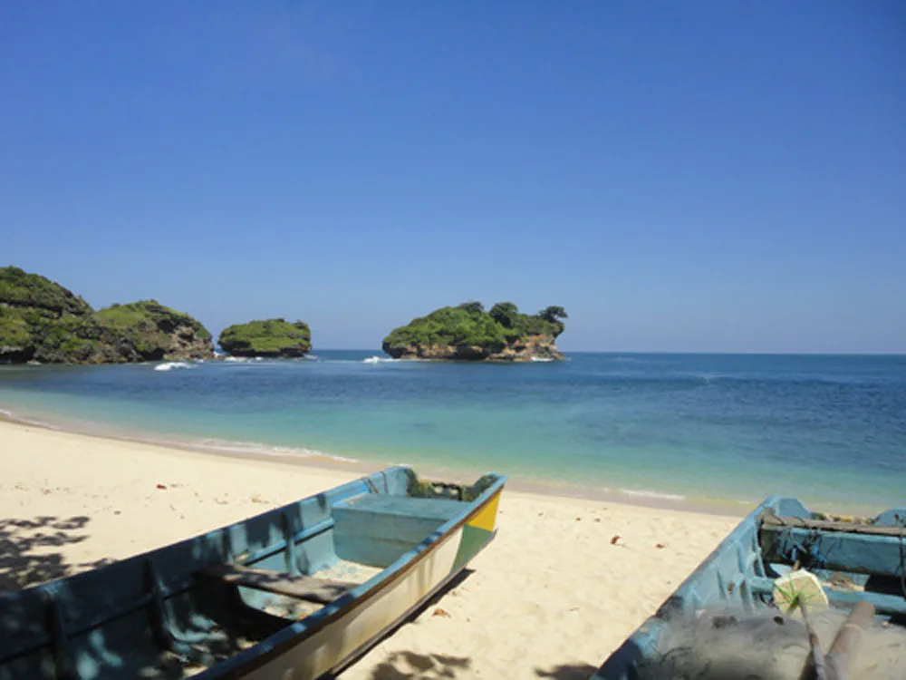 Dua perahu kayu nelayan bersandar di pasir putih Pantai Watu Karung dengan tiga pulau karang bertopi hijau di lepas pantai