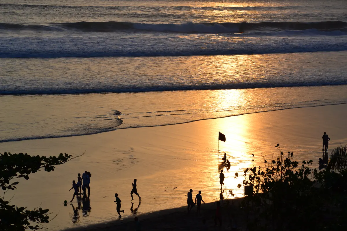 Pengunjung bermain di garis ombak Pantai Seminyak saat cahaya matahari terbenam memantul keemasan di pasir basah