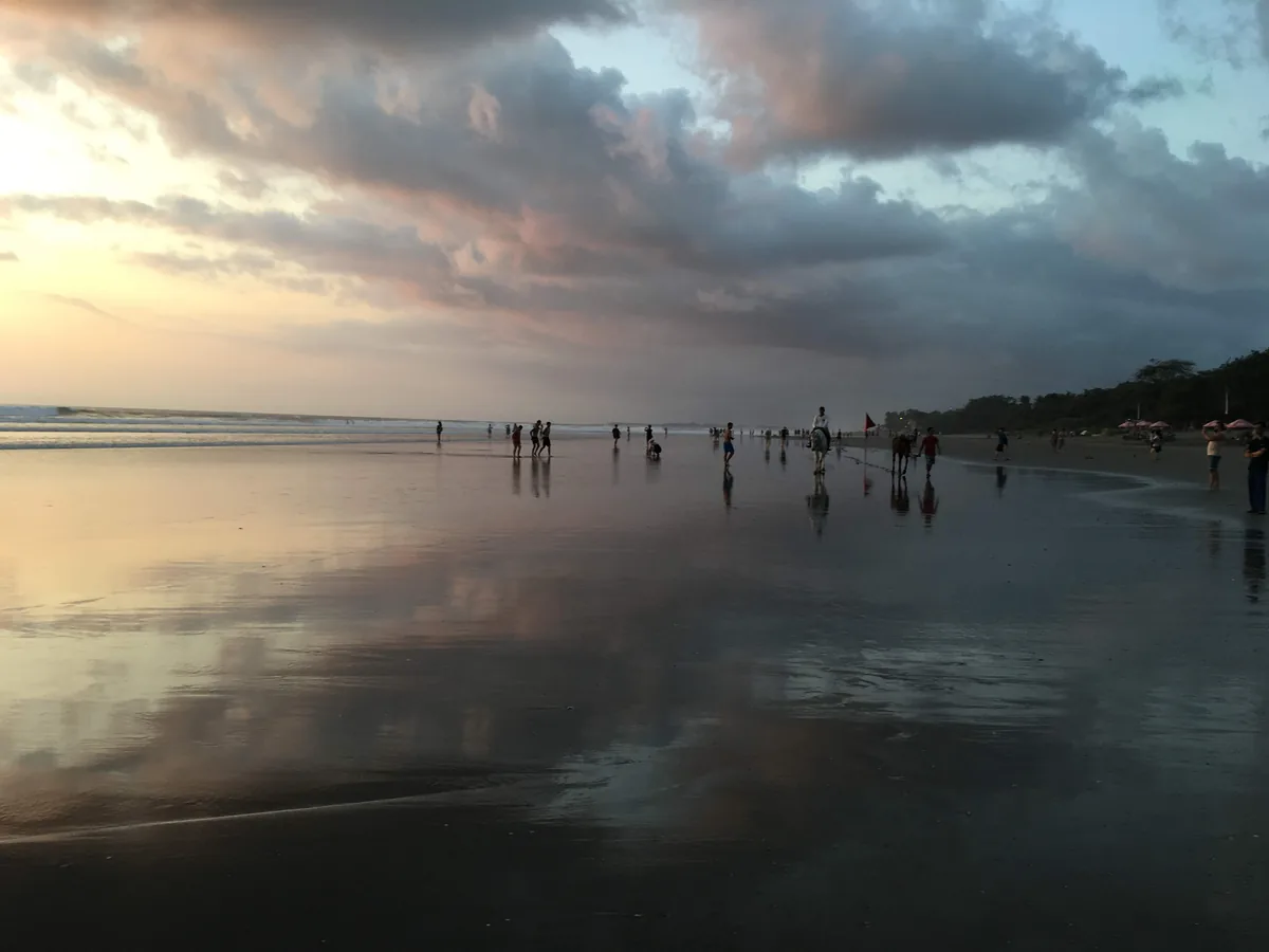 Hamparan pasir basah Pantai Seminyak memantulkan awan senja, dengan pejalan kaki dan penunggang kuda di kejauhan