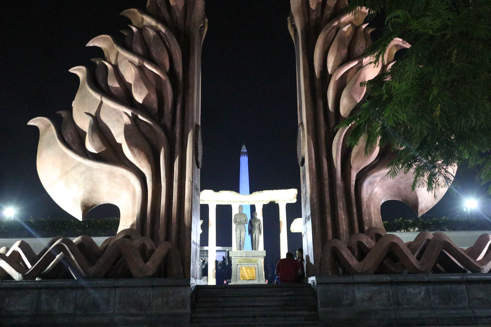 Patung Soekarno dan Hatta di bawah gapura berbentuk lidah api dengan obelisk Tugu Pahlawan menjulang di belakangnya pada malam hari