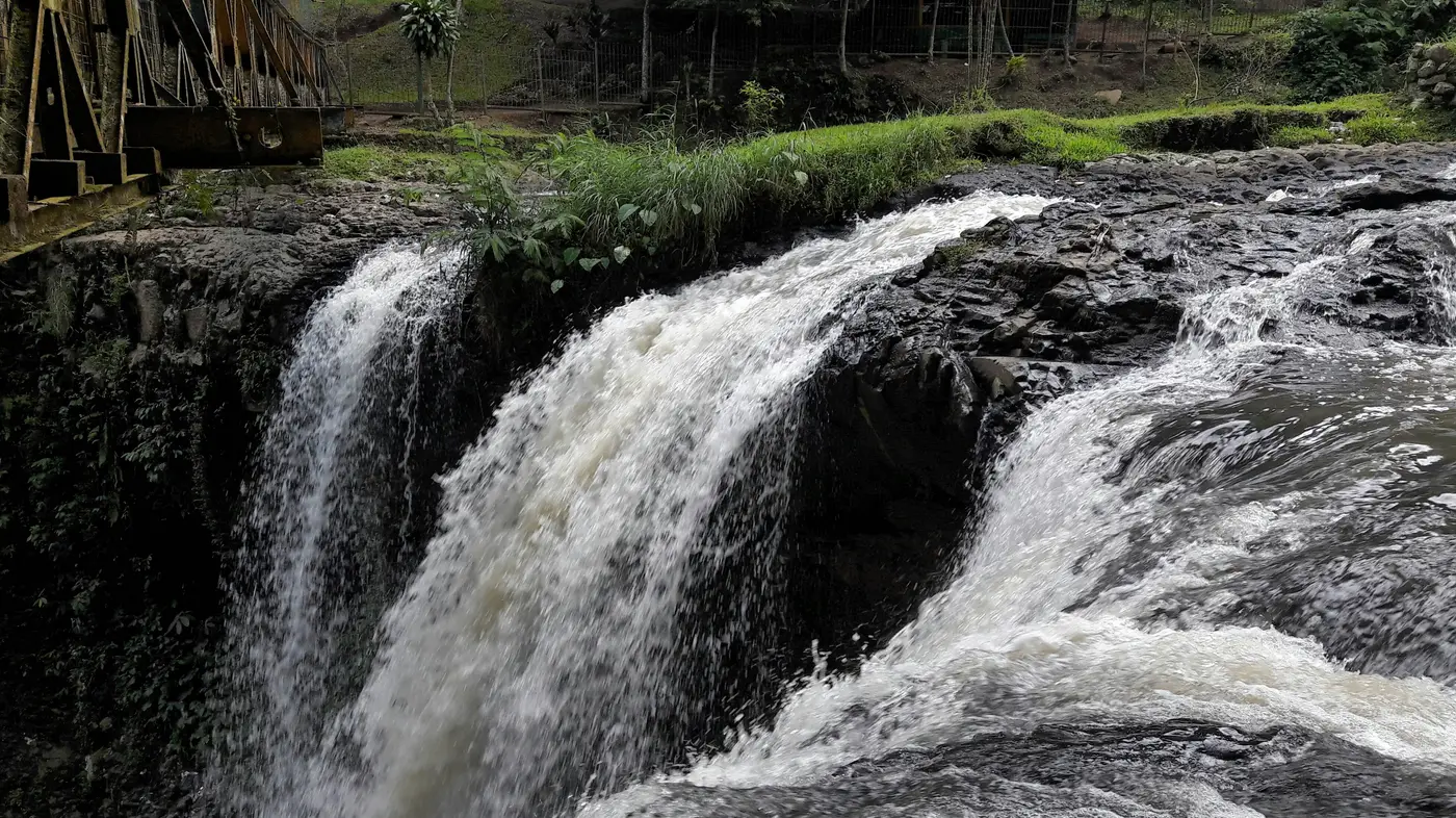 Air Terjun Curug Omas mengalir deras di atas batuan gelap dengan rangka jembatan besi di sisi kiri atas