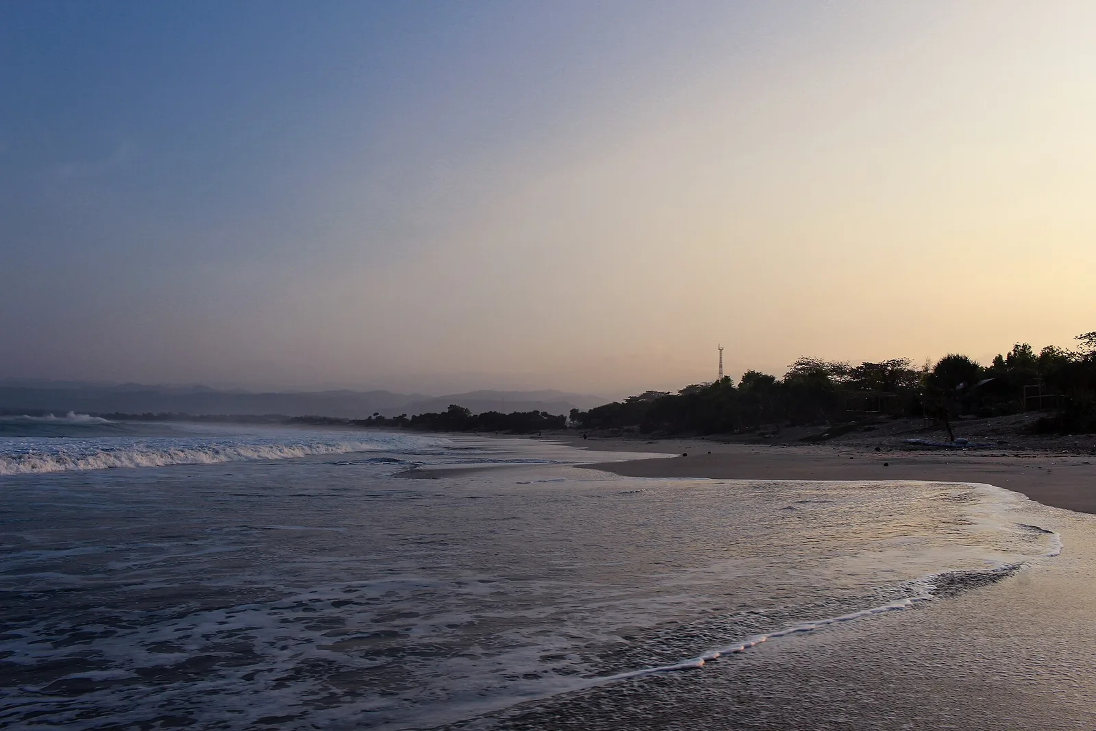 Garis Pantai Santolo di Garut selatan saat senja, ombak putih bergulung menyapu pasir dengan perbukitan samar di kejauhan